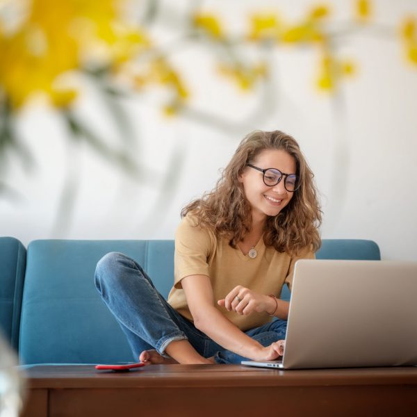 Charming beautiful young girl in glasses with curly hair sits on a blue sofa at home in front of a laptop, remote work and education, beauty and fashion.